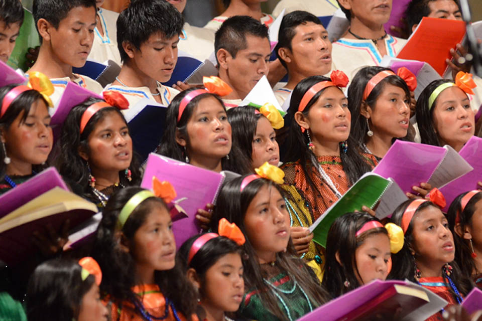 A group of Bolivian singers, with female singers wearing headbands with bright orange and yellow flowers on them on their dark hair, and male singers in white shirts, performing as a group.